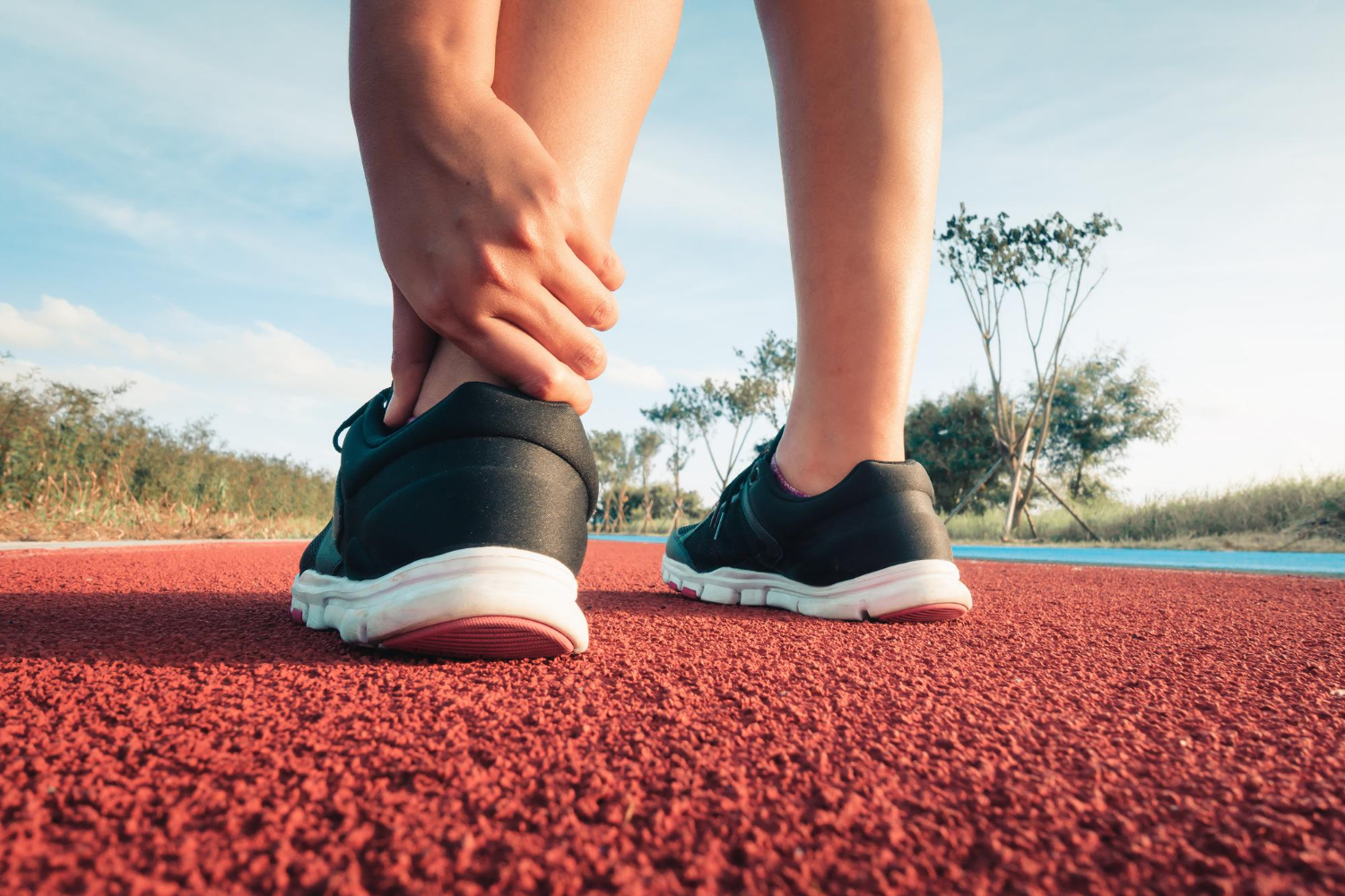 Close-up of a person adjusting their left ankle in black running shoes on a red outdoor track.