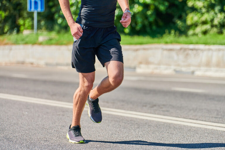 Man running on a paved road in black athletic shorts and gray running shoes, mid-stride on a sunny day.