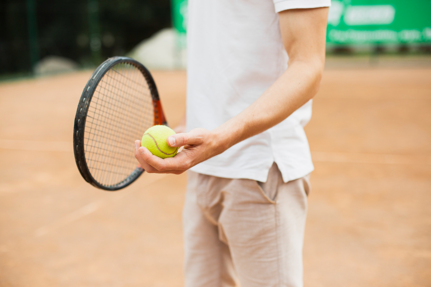 Person on a tennis court holds a bright yellow tennis ball next to a racket, ready to play on a clay surface.